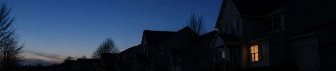 A dark suburban street at dusk during a blackout, with one window lit by lantern light
