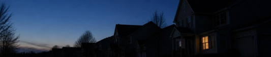 A dark suburban street at dusk during a blackout, with one window lit by lantern light
